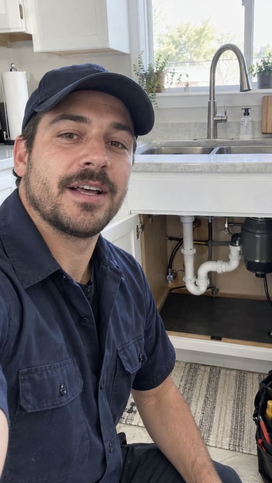 Raw, unedited phone video frame of a plumber giving a service call update in front of an open kitchen sink cabinet.